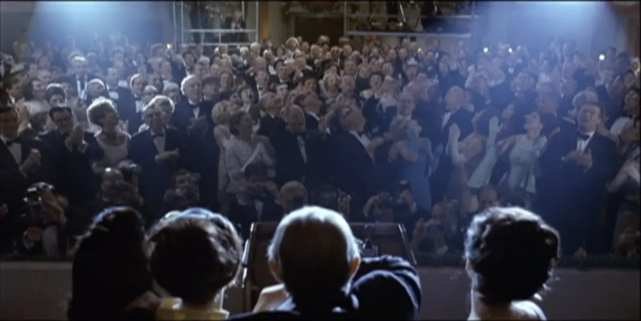 Path to War (2003) movie still: wide — A crowded auditorium filled with people in formal attire faces a speaker on stage. The audience stands and…