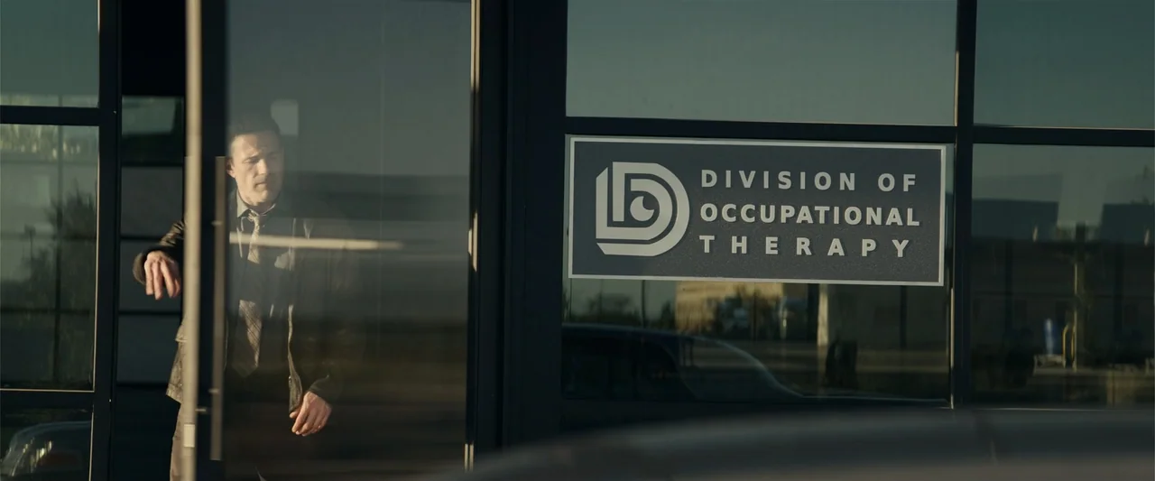 Hypnotic (2023) movie still: medium — A glass exterior wall of a commercial building features a sign for occupational therapy. A man in a dark…