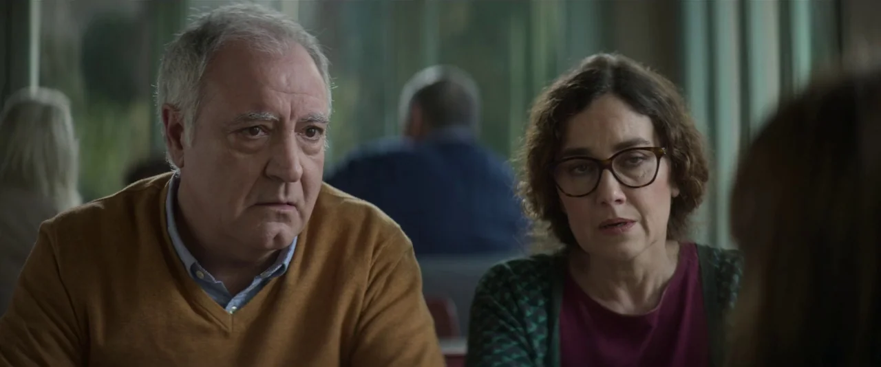 Crazy About Her (2021) movie still: medium — An older couple sits together in a booth inside a restaurant setting. They look ahead at a person…