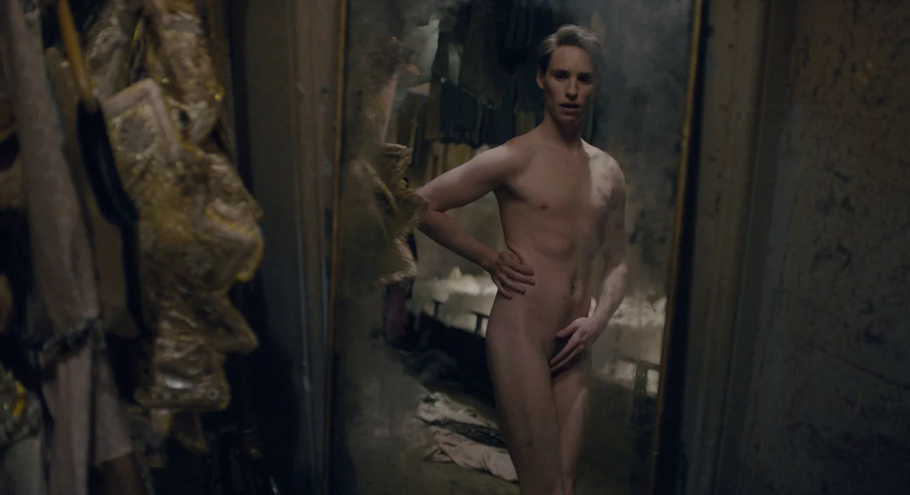 The Danish Girl (2015) movie still: fullbody — A dimly lit room features a large, vintage mirror filled with ornate fabric clutter. A man stands…