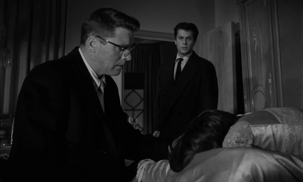 Sweet Smell of Success (1957) movie still: medium — A dimly lit room contains a bed with a person lying down in the foreground. One man in glasses…