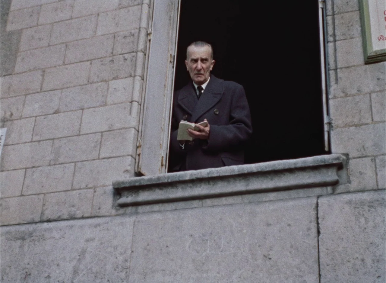 The Red Balloon (1956) movie still: medium — An aging man stands at a stone window opening of a light-colored building. He holds a small,…