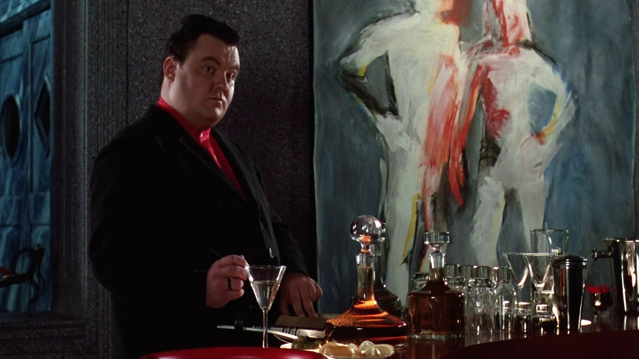 Beetlejuice (1988) movie still: medium — A modern bar area features a large abstract painting of two figures behind him. A man in a suit holds a…