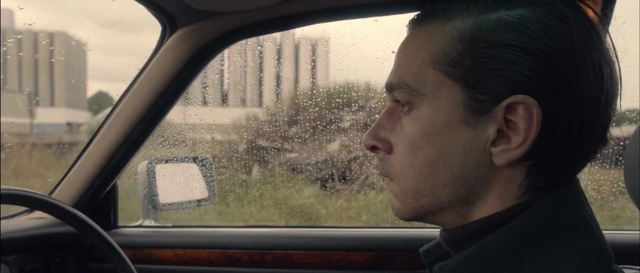 Nymphomaniac Vol 2 (2013) movie still: medium — A man sits in the driver seat of a car during a light rain. The window is covered in raindrops,…