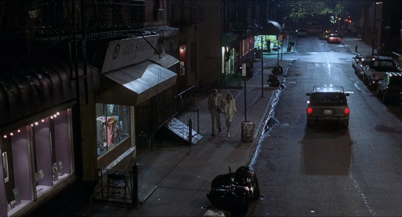 The Prince of Tides (1991) movie still: wide — A nighttime city sidewalk passes by a storefront with illuminated windows and trash bags. A couple…