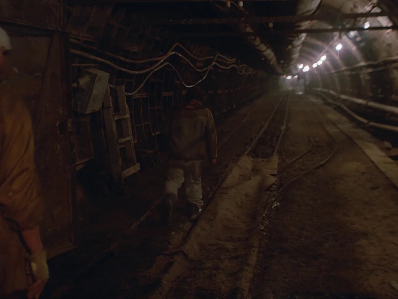 Kin-dza-dza! (1986) movie still: fullbody — A dimly lit industrial mine tunnel stretches deep into the darkness along metal tracks. One man walks…