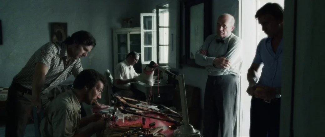 Munich (2005) movie still: wide — A dimly lit room functions as a makeshift operations center with minimal furniture. Five men stand or sit around a…