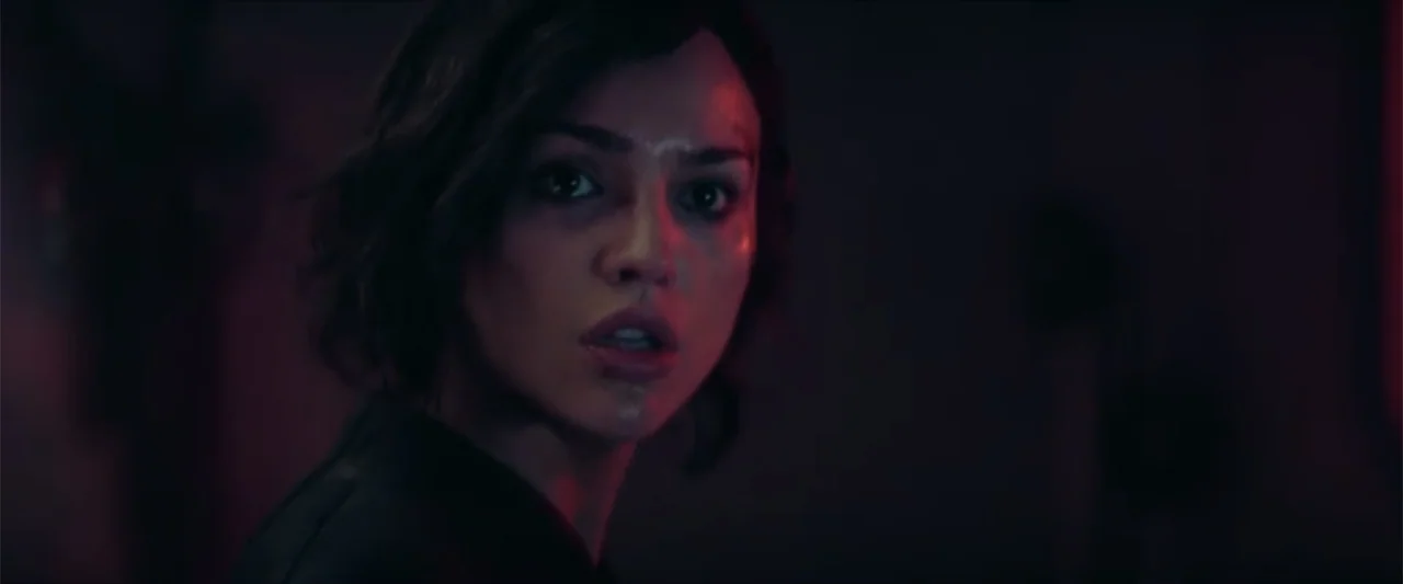 Ash (2025) movie still: closeup — A dark interior room is bathed in low-key red lighting. A young woman with dark hair looks toward the side of the…