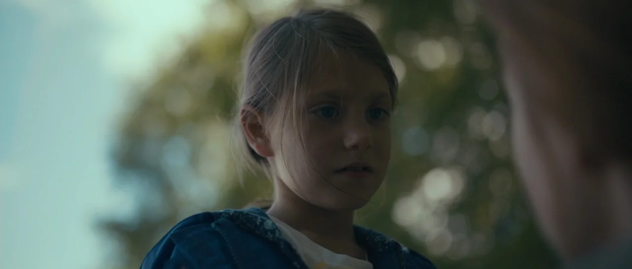 Arrival (2016) movie still: closeup — A child stands outdoors against a background of blurred green trees. The girl looks down and away from the…