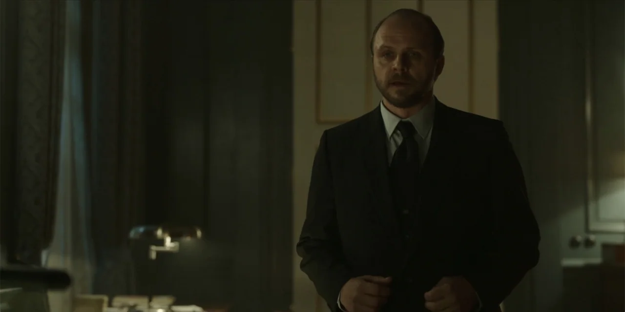 Oslo (2021) movie still: medium — A dim professional office features dark wood paneling and subtle classic architectural trim. A man in a dark…