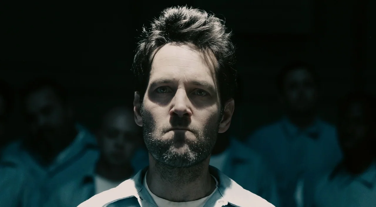 Ant-Man (2015) movie still: closeup — A man stands in a dark space surrounded by out-of-focus figures in prison uniforms. He looks directly toward…
