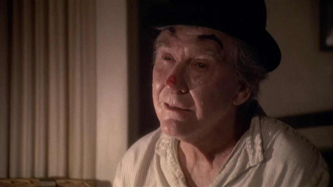 The Day of the Locust (1975) movie still: closeup — A dimly lit room with plain walls features an elderly man. He wears a bowler hat and light clown…