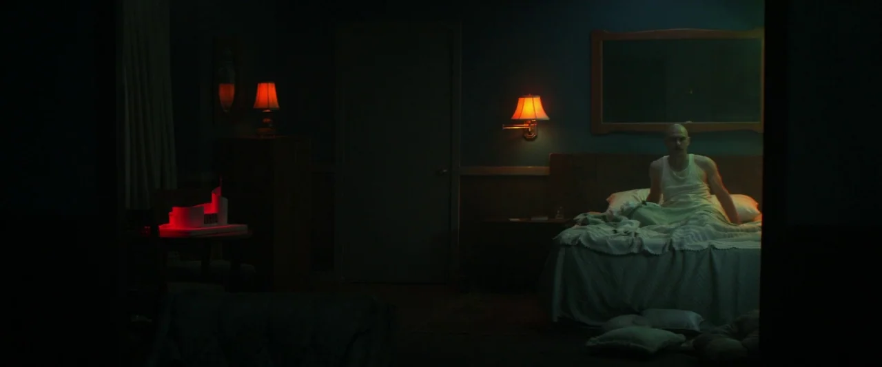 Zeroville (2019) movie still: wide — A dimly lit motel room features a man sitting in bed at night. A red glowing model structure sits on a small…
