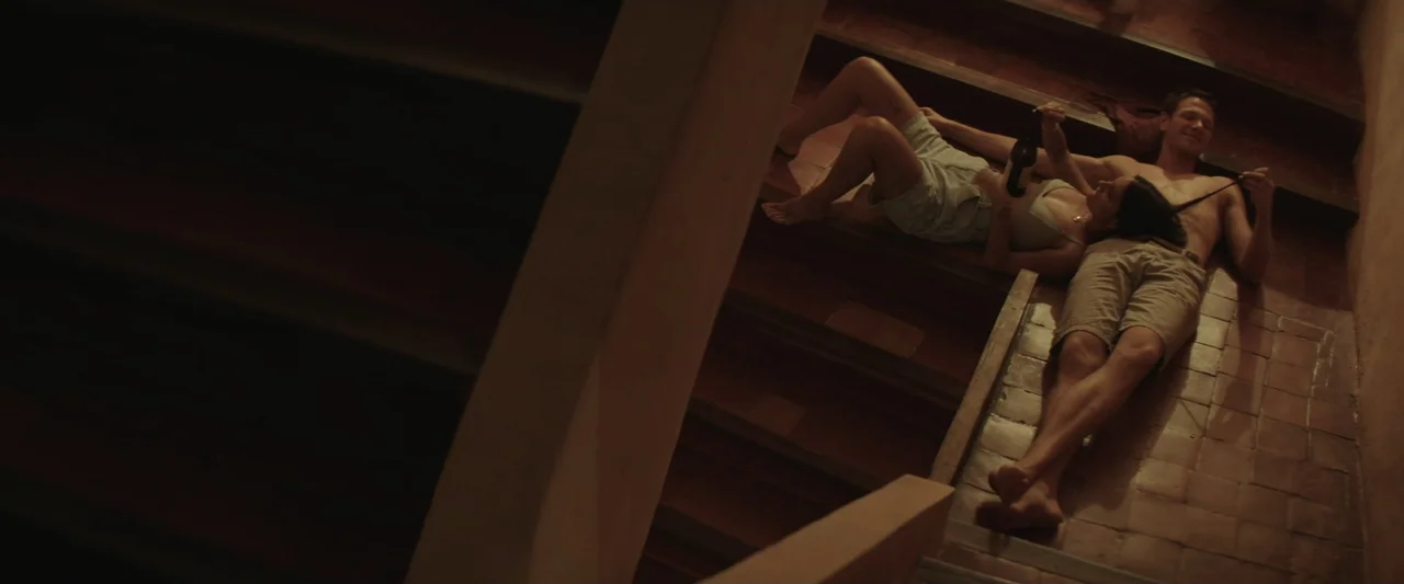 The Only One (2021) movie still: wide — A dimly lit tiled staircase inside a building with dark wood accents. A man and woman lie together on the…