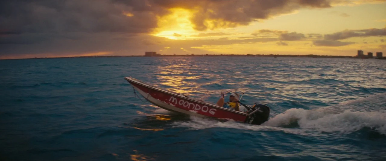 The Beach Bum (2019) movie still: wide — A small motorboat with painted lettering traverses open water during a golden sunset. A lone man sits in…