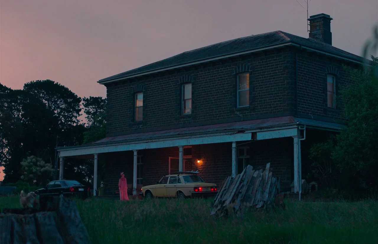 Nitram (2021) movie still: wide — A two-story brick farmhouse sits in a field during the fading twilight hours. A woman in a pink dress stands near…