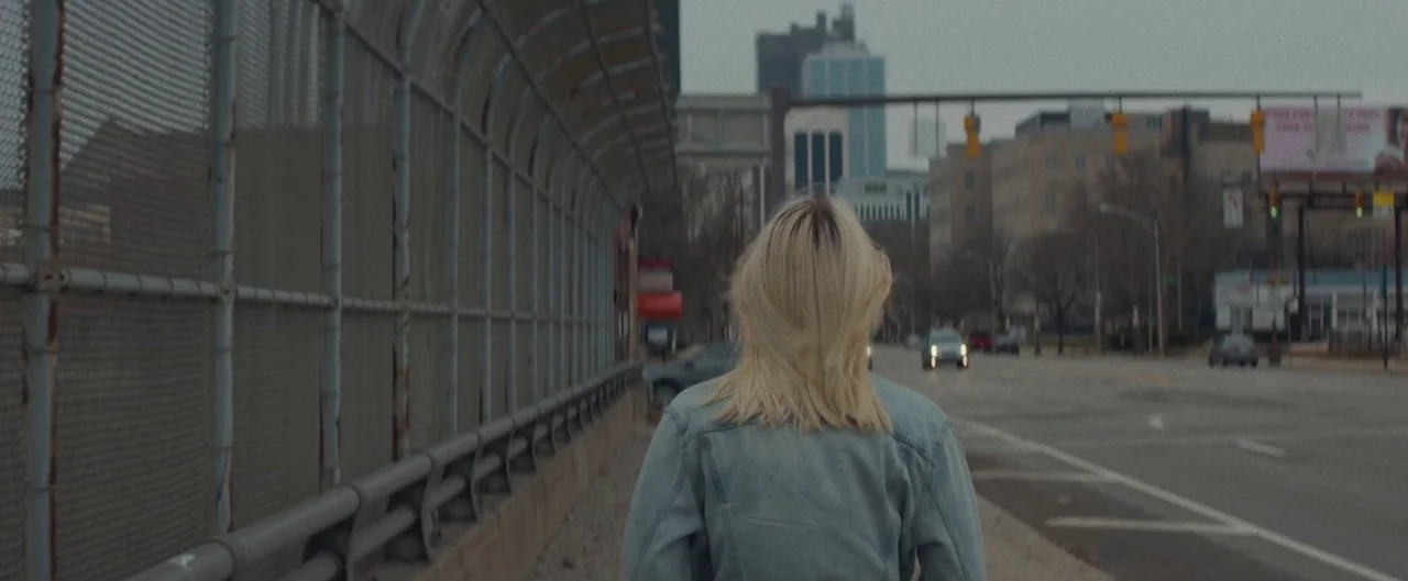 Poser (2021) movie still: medium — A woman walks along a paved city sidewalk bordered by a tall chain-link fence. A multi-lane road and downtown…