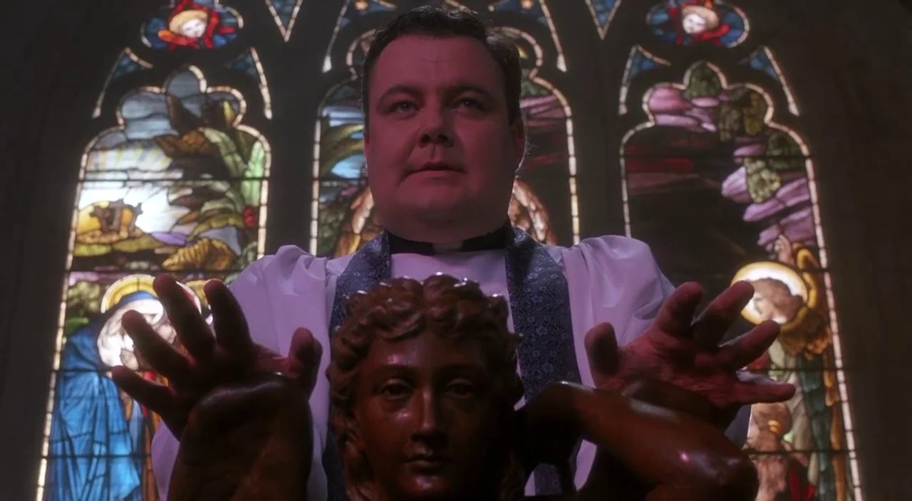 Heathers movie still: closeup — A man stands before a multi-pane stained glass window depicting religious figures in a church. The priest holds both…