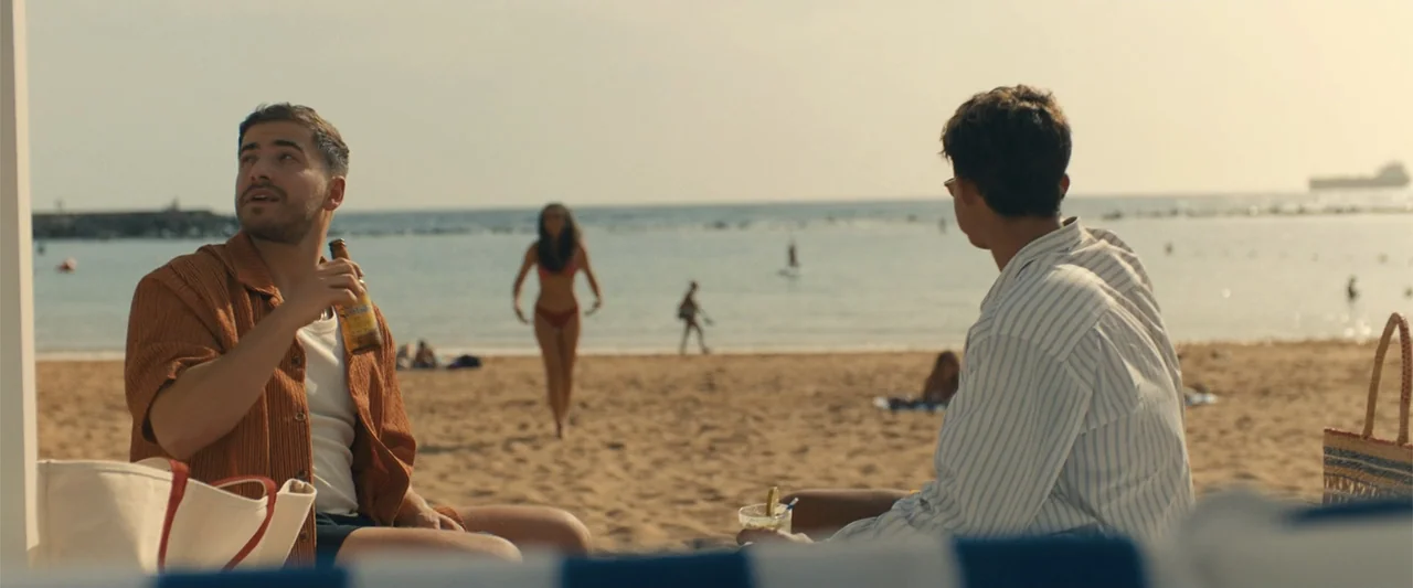 My Fault: London (2025) movie still: medium — A sunny public beach with scattered people in the background during golden hour. Two men sit in the…
