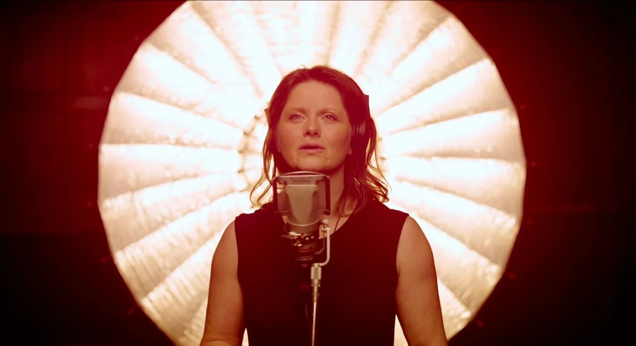One More Time With Feeling movie still: medium — A woman stands in front of a bright circular light in a dark space. She sings into a metallic…