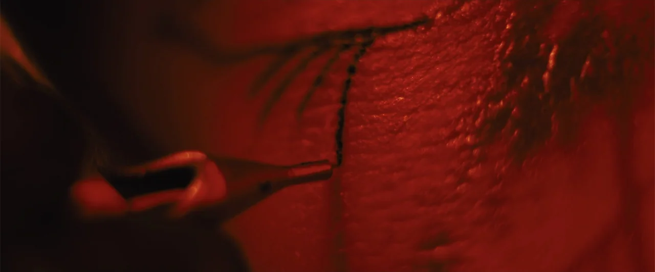 Skin (2009) movie still: closeup — Close-up view of a tattoo machine needle piercing human skin with black ink. Red lighting bathes the entire frame…