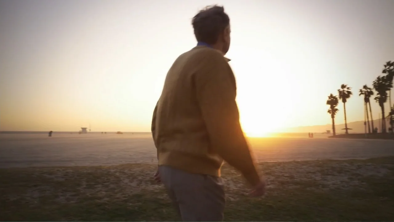 Beware of Angels (2016) movie still: medium — A wide sandy beach at sunset features tall palm trees against the bright sky. A man in a yellow…