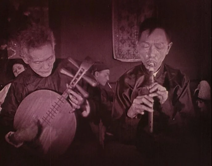 Broken Blossoms (1919) movie still: medium — A dimly lit room features two men playing traditional musical instruments. A person sits partially…