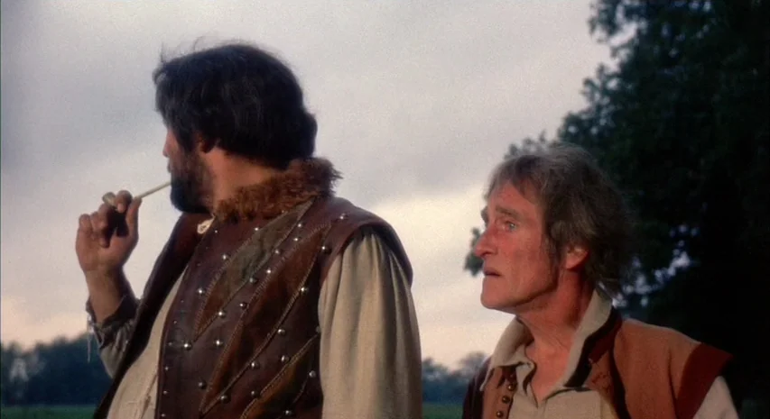 Witchfinder General (1968) movie still: medium — Two men stand outdoors in an open field against an overcast sky. The man on the left smokes a pipe…