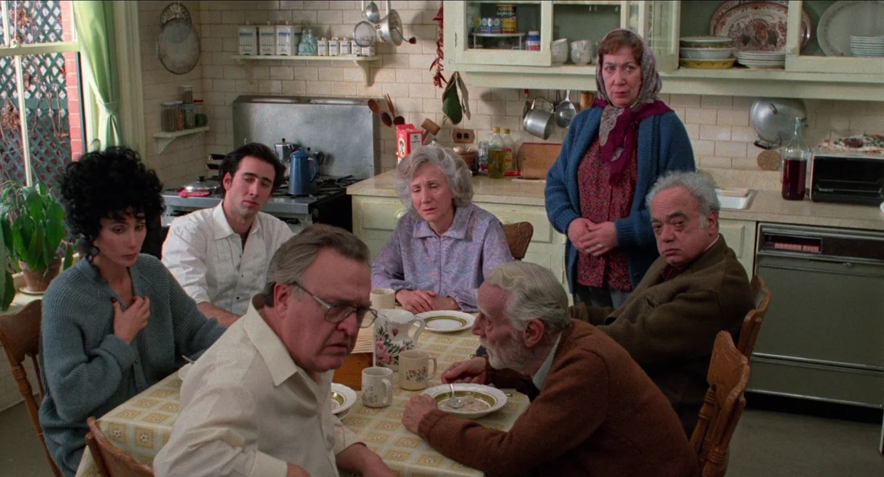 Moonstruck (1987) movie still: medium — A crowded kitchen with cream-colored cabinetry and tile walls during the day. A group of six people sits…