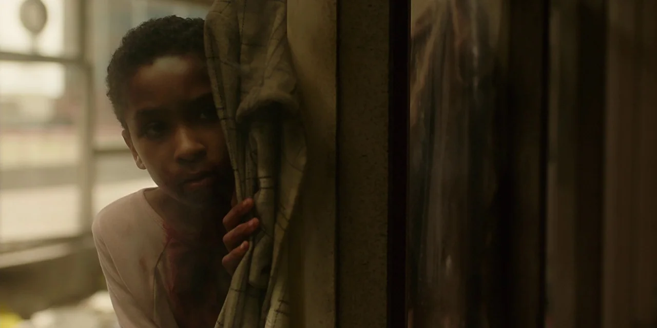 The Girl With All The Gifts (2016) movie still: closeup — An interior space features a child partially hidden behind a light-colored fabric curtain.…