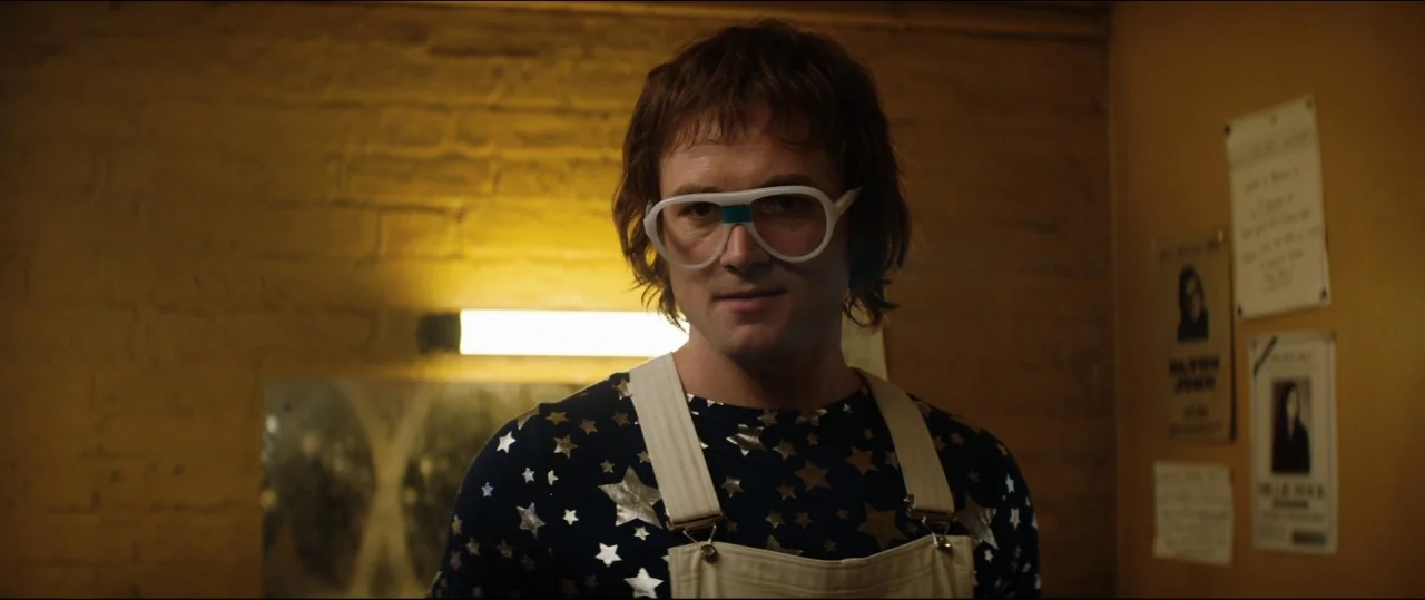 Rocketman (2019) movie still: medium — A dimly lit room with textured yellow brick walls and paper flyers pinned up. A man wearing white-framed…