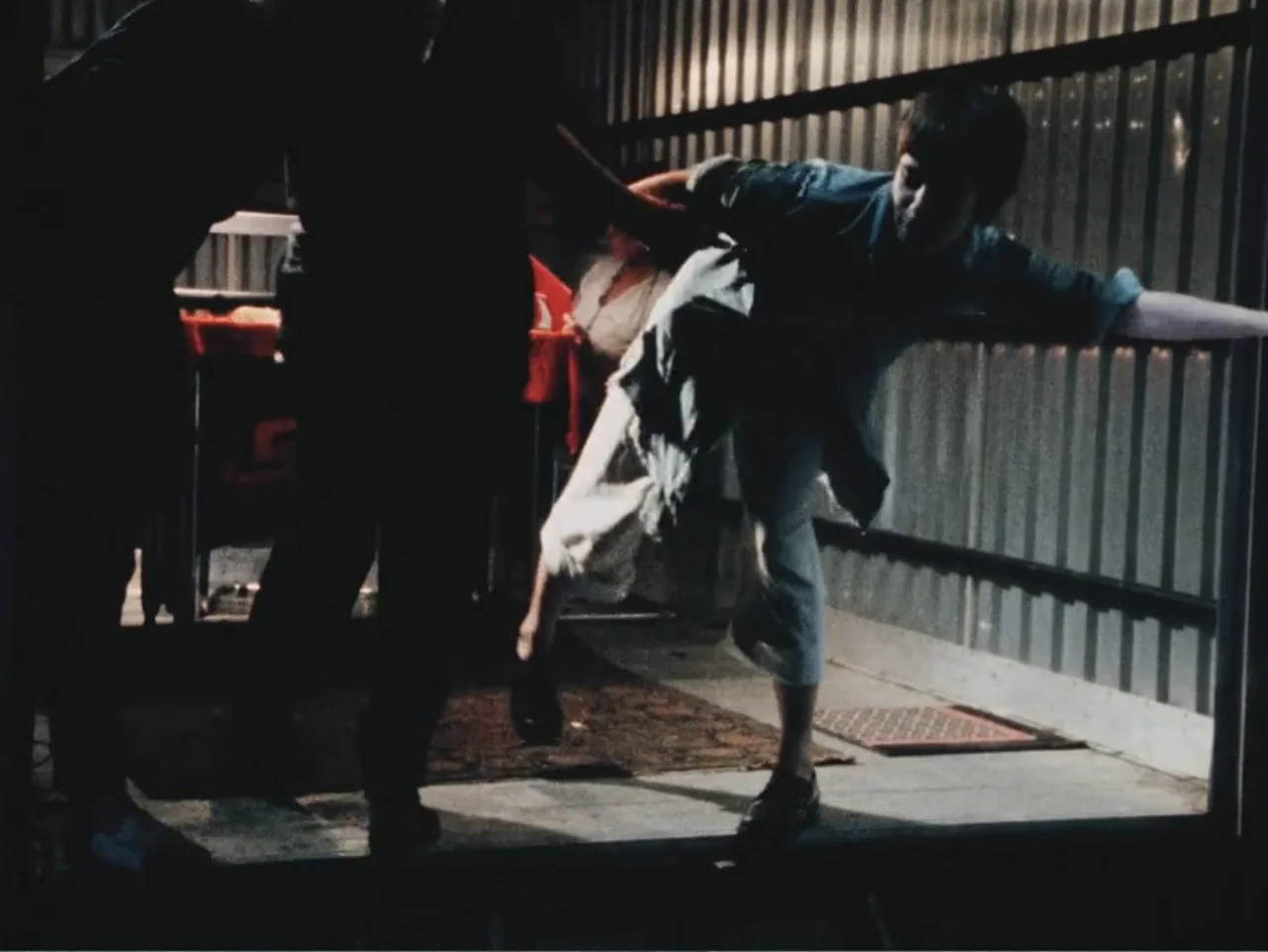 Born in Flames (1983) movie still: medium — A dimly lit industrial space with corrugated metal walls and concrete floor. A person walks forward from…