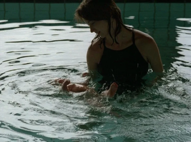 Hail Mary (1985) movie still: medium — An indoor swimming pool with blue tiled walls is filled with water. A woman wearing a black swimsuit holds a…