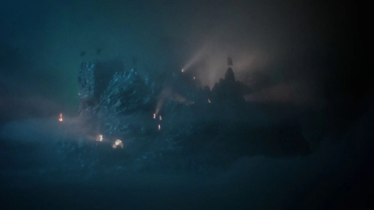 Excalibur (1981) movie still: establishing — A stone fortress sits atop a jagged rock formation surrounded by thick dark fog. Several small fires…