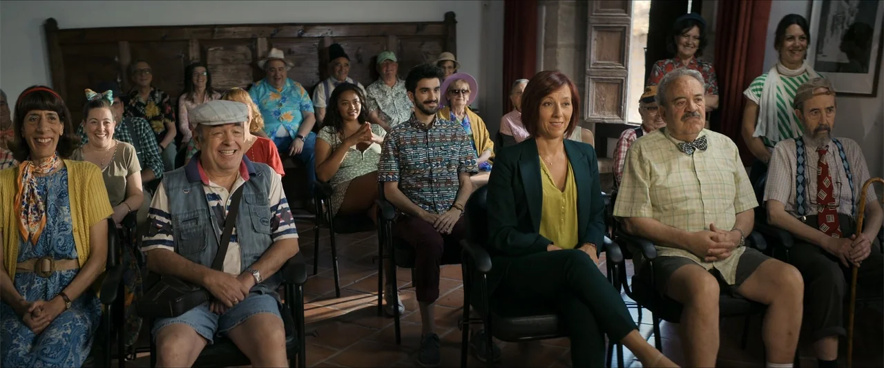A Hipster in Rural Spain (2024) movie still: medium — A group of diverse adults sit in chairs arranged in rows inside a hall. The subjects face…