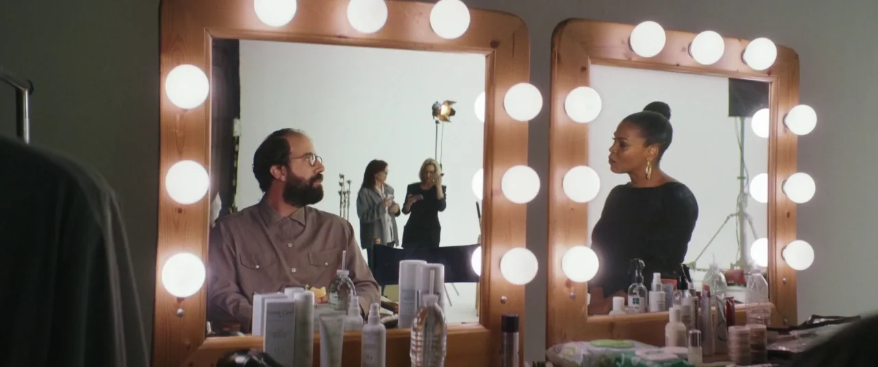 Lemon (2017) movie still: medium — Two light-framed vanity mirrors are mounted on a white wall in a studio space. A man and a woman sit before their…