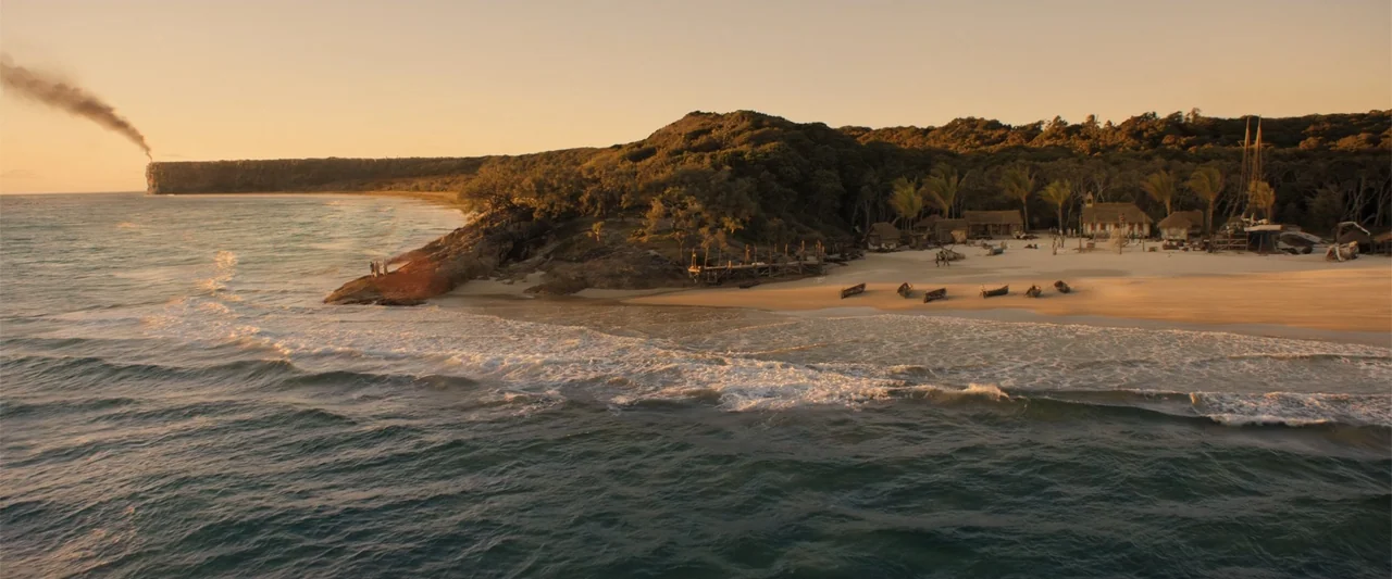 The Bluff (2026) movie still: establishing — A coastal settlement sits on a sandy beach below a dense treeline and cliff. Small wooden boats line…