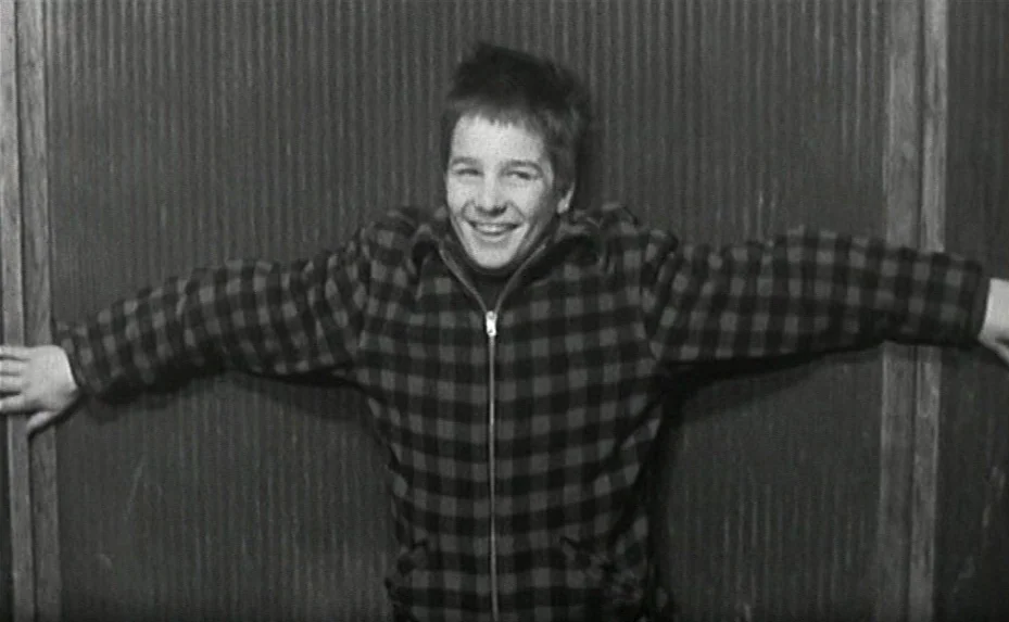 Love on the Run (1979) movie still: medium — A teenage boy stands against a ribbed, dark wall in a narrow corridor. He smiles while holding his arms…