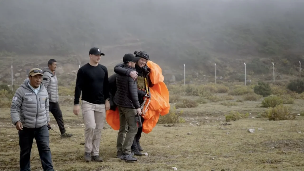 Drop Zone Everest (2023) movie still: fullbody — An overcast mountainous field with sparse vegetation and a distant wire perimeter fence. A group of…