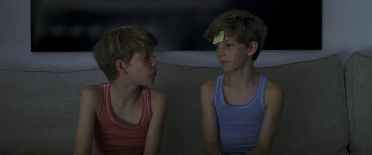Goodnight Mommy (2014) movie still: medium — Two boys sit on a beige couch against a plain wall. One boy has a yellow sticky note attached to his…