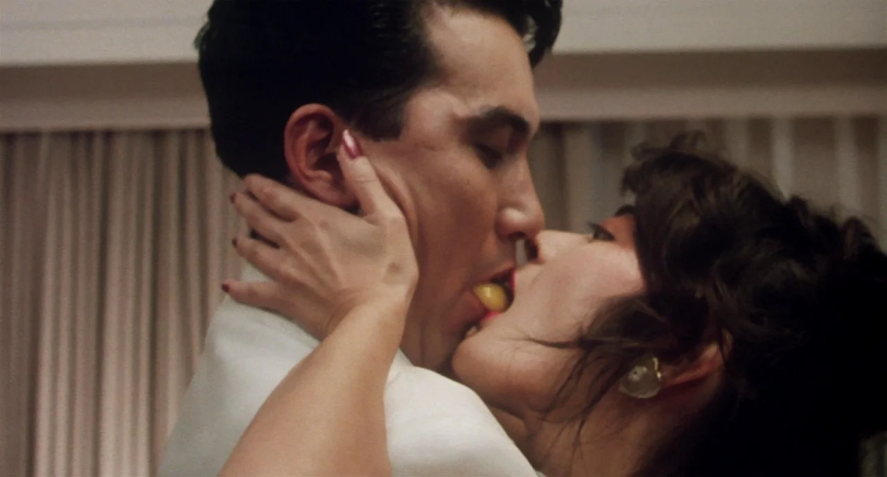 Tampopo (1985) movie still: closeup — A man and woman are embracing closely in a room with light curtains. The woman holds a grape in her mouth as…