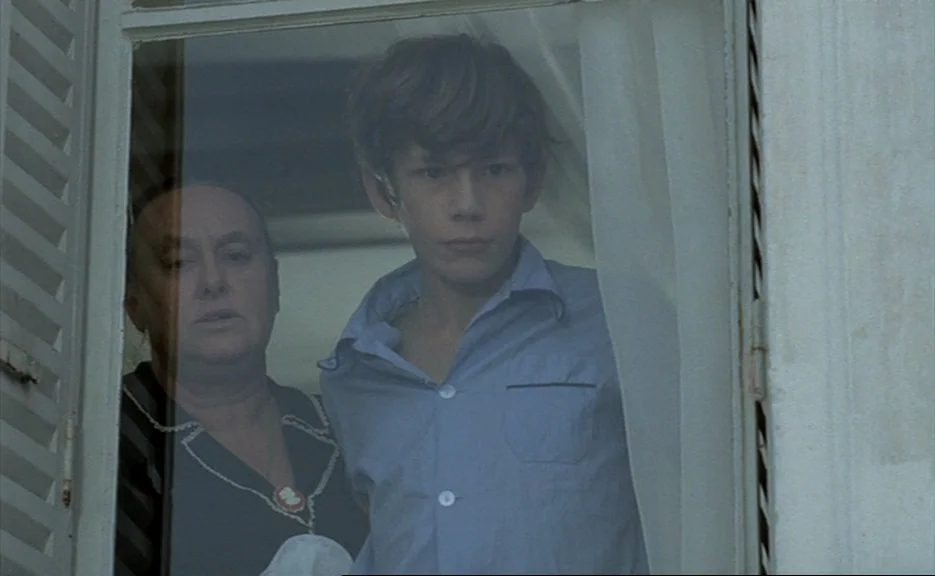 Murmur of the Heart (1971) movie still: medium — An older woman and a young boy stand behind a white wooden window frame. They look outward through…