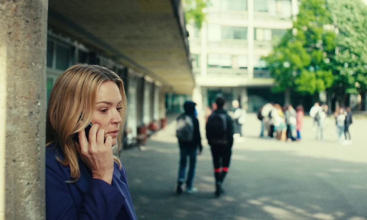 Just The Two Of Us (2023) movie still: medium — An outdoor paved area in front of a modern multi-story concrete educational building. A woman stands…