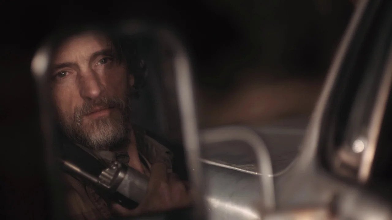 Winter’s Bone (2010) movie still: closeup — The face of a middle-aged man is visible in the reflection of a side mirror. He holds a firearm while…