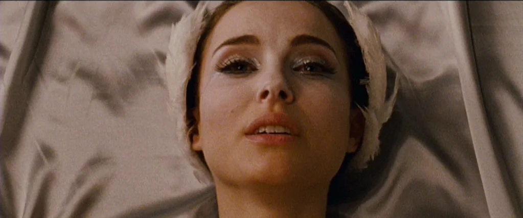 Black Swan (2010) movie still: closeup — A woman lies on a bed with light-colored, reflective satin bedding. She wears a white feather headpiece and…