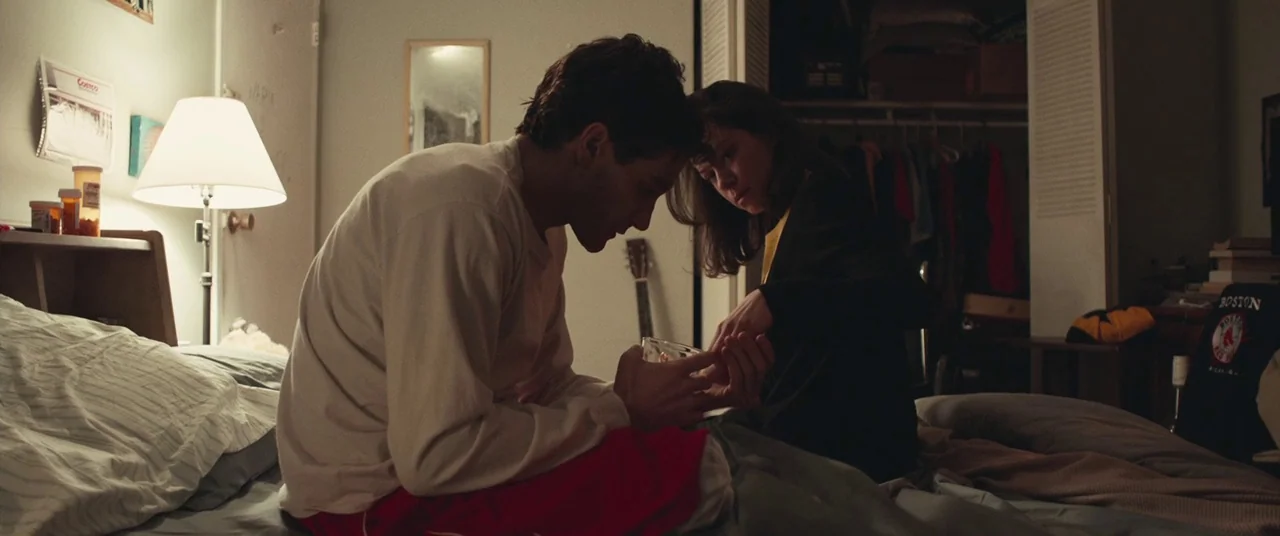 Stronger (2017) movie still: medium — A dimly lit room with an unmade bed and a cluttered nightstand. A woman interacts with a man sitting on the…
