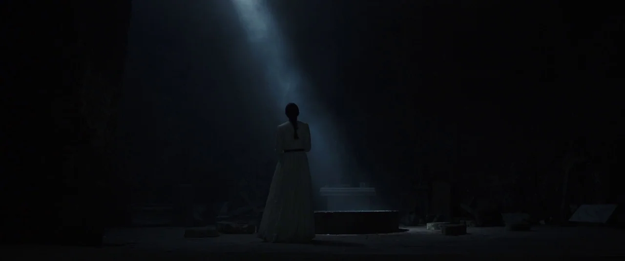 The Nun (2018) movie still: fullbody — A dark, subterranean stone chamber features a single beam of light piercing the gloom. A woman in a white…