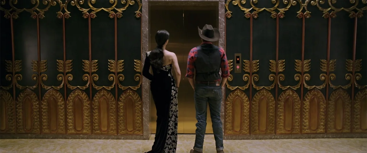 Skiptrace (2016) movie still: fullbody — Two individuals stand in front of decorative dark green and gold elevator doors. The woman wears a formal…