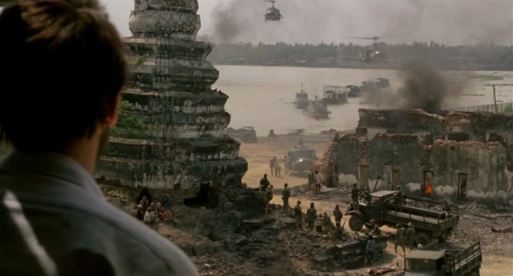 The Killing Fields (1984) movie still: wide — A riverside encampment features a crumbling stone pagoda and military vehicles near smoke. Soldiers…