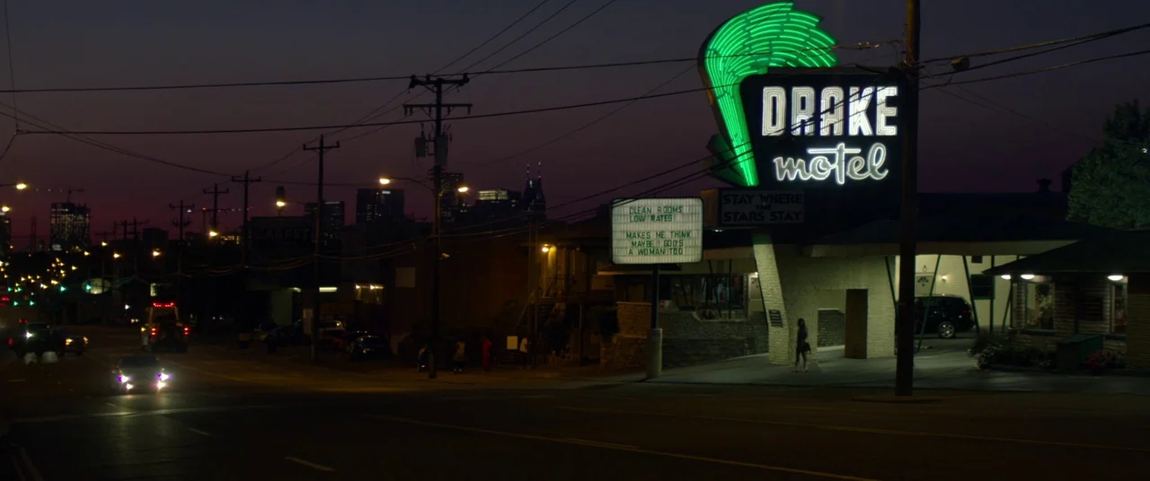 Wild Rose (2018) movie still: wide — A motel at night features a large, bright neon sign and a message board. The parking lot sits alongside a dark…
